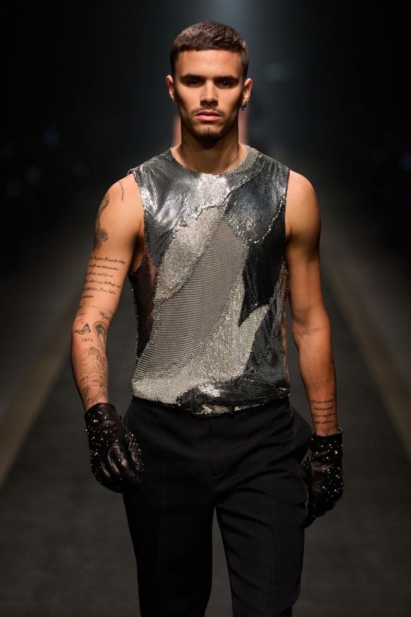 Romeo Beckham walking down the runway wearing a metallic mesh sleeveless top, black trousers, studded black gloves, and displaying his tattooed arms.