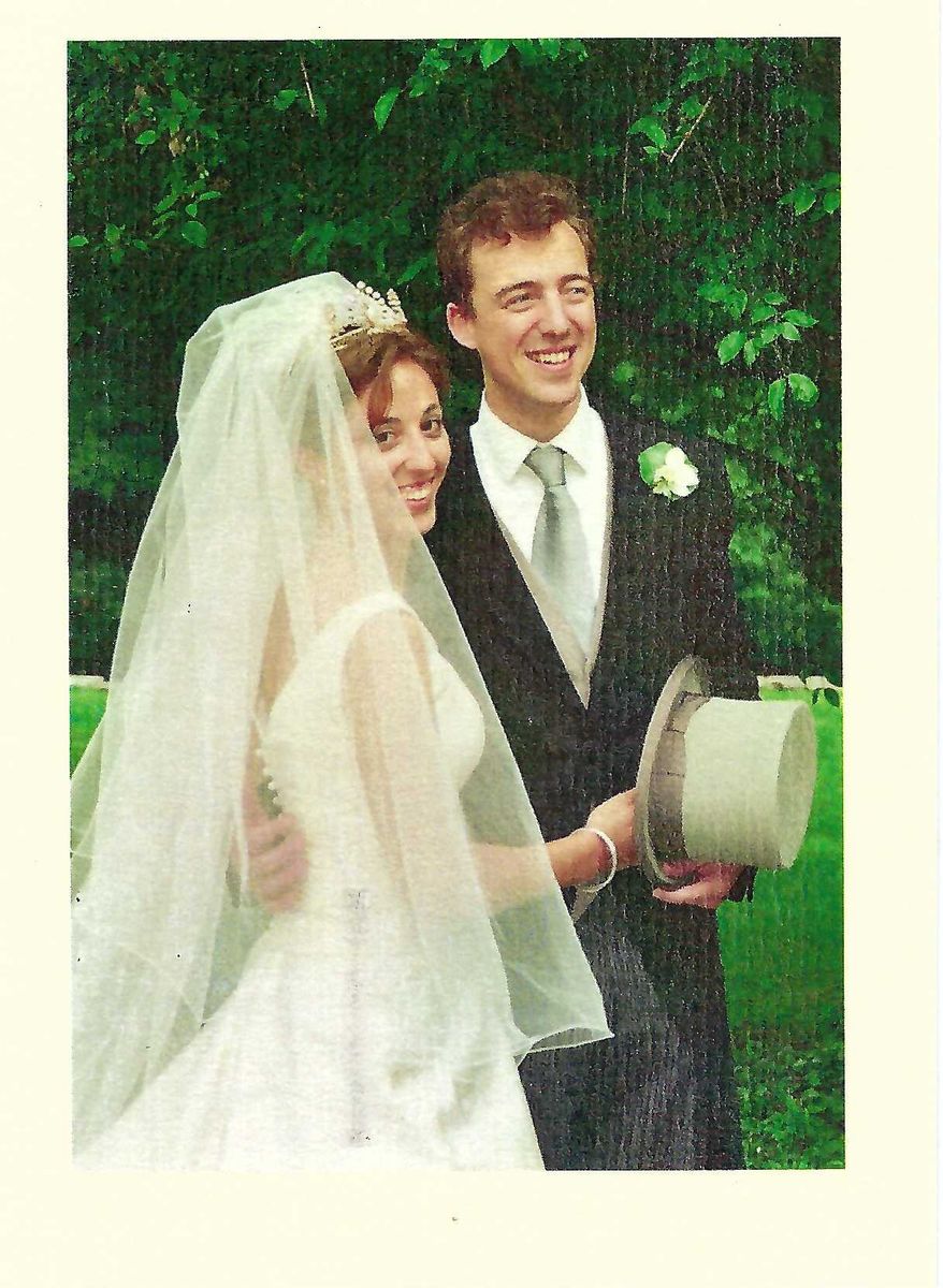 A smiling bride and groom, with the bride wearing a tiara and veil, and the groom in a formal suit holding a top hat, posing outdoors on their wedding day.