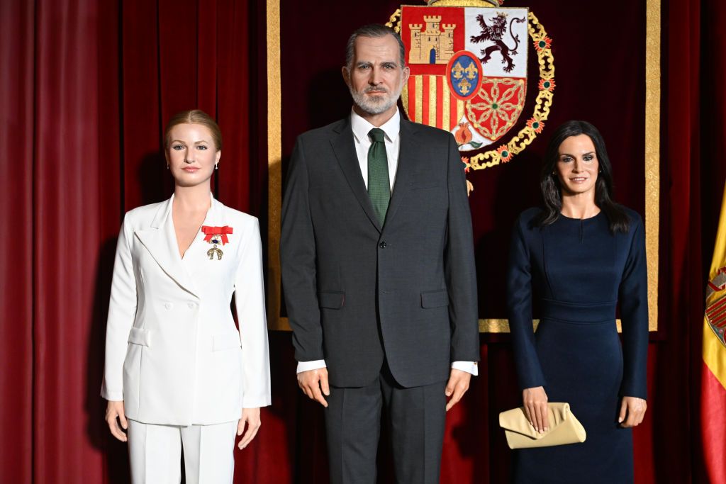 Wax figures of Princess Leonor, King Felipe VI, and Queen Letizia stand side by side at the Madrid Wax Museum, with the Spanish coat of arms and flag in the background.