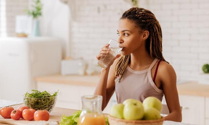 Woman drinking water in her kitchen with healthy foods around her