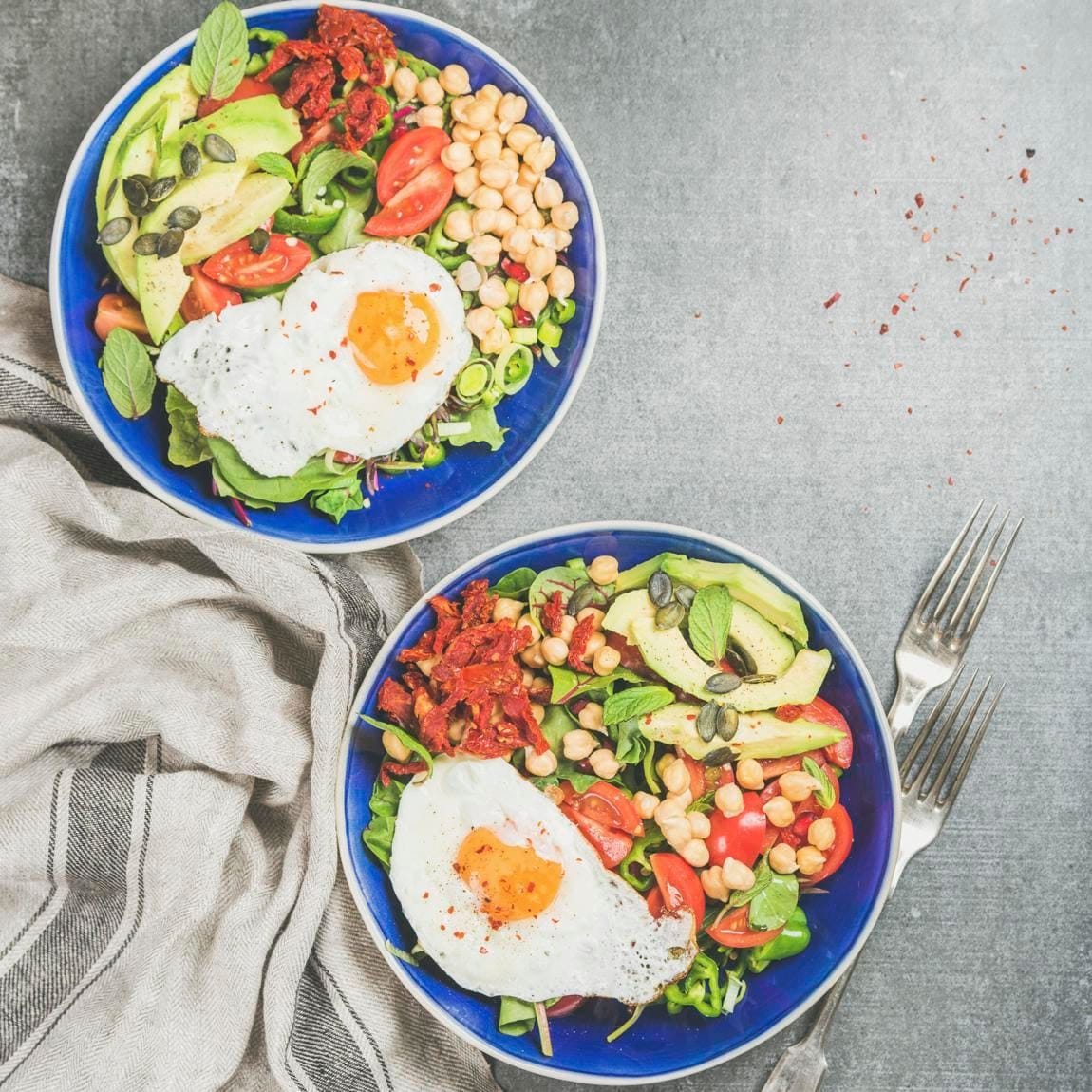 Healthy breakfast bowls with fried egg, chickpea sprouts, seeds, vegetables and greens over grey concrete background