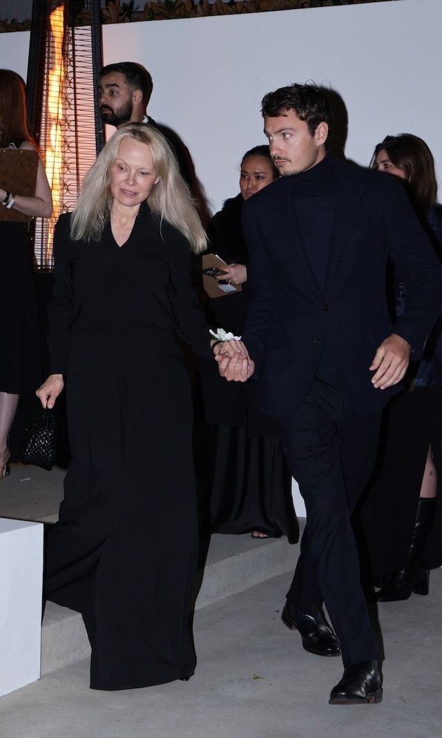 Pamela Anderson walks hand in hand with a companion at a pre-Oscar event, wearing a black floor-length dress with a deep V-neckline and long sleeves. She keeps her look simple with minimal makeup and flowing blonde hair, exuding natural beauty and sophistication.