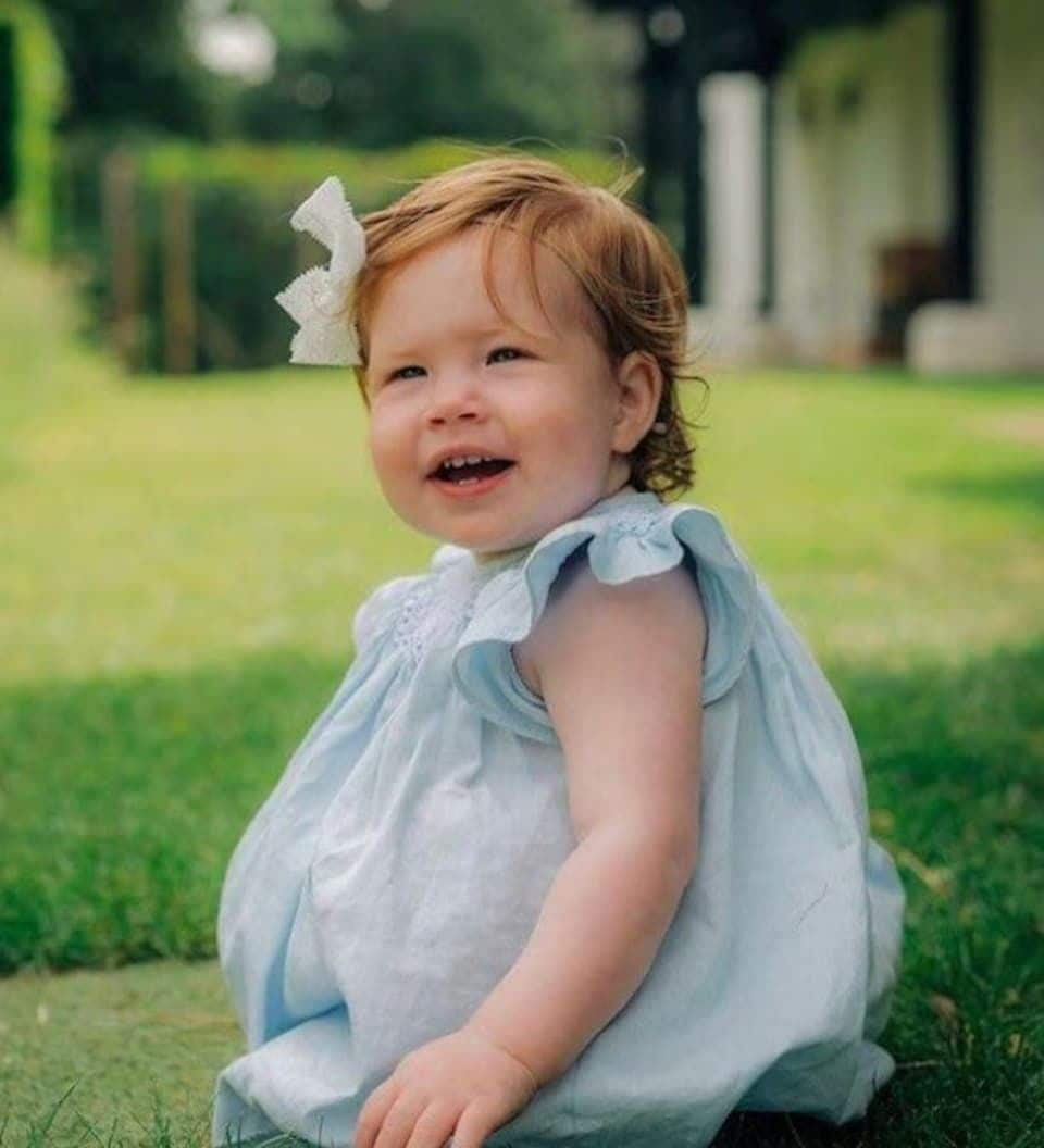 Lilibet Diana, a young child with red hair, smiles while sitting on the grass in a light blue dress with a white bow in her hair.