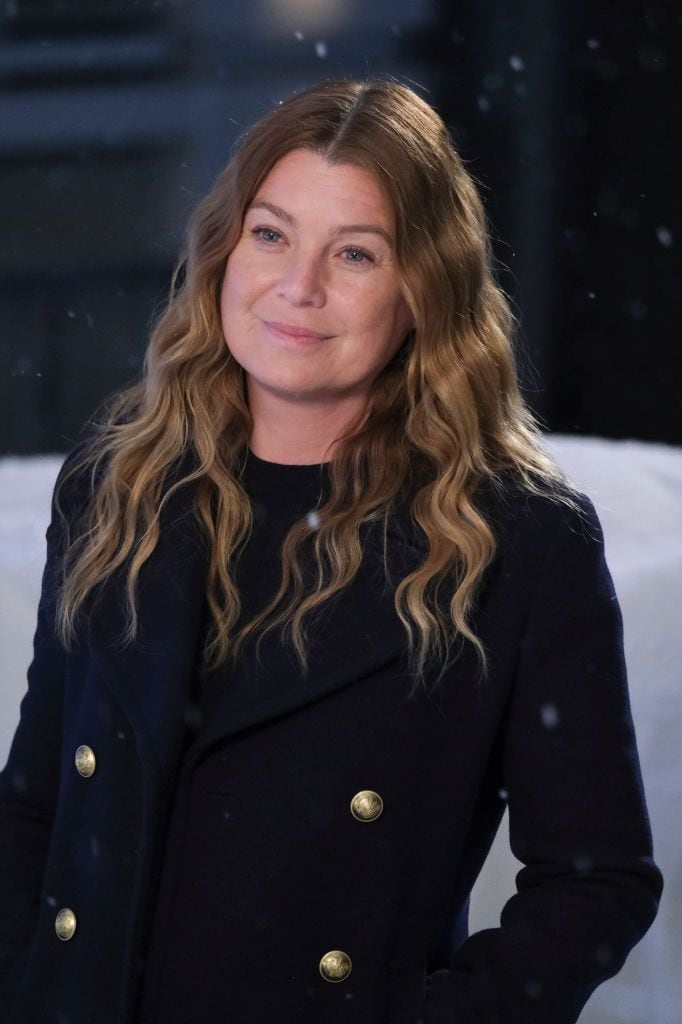 Ellen Pompeo en un abrigo azul marino con botones de oro, parado al aire libre en un entorno nevado, sonriendo suavemente.