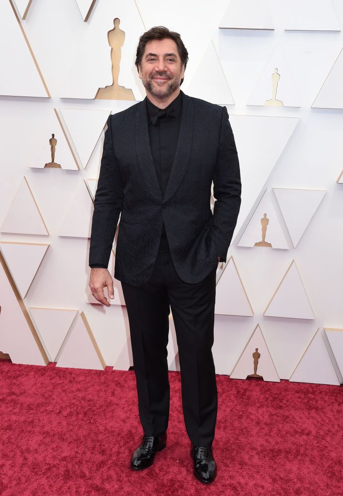 Javier Bardem poses on the red carpet at the 2024 Oscars, wearing a stylish black suit with a subtle pattern, smiling confidently against the event backdrop.