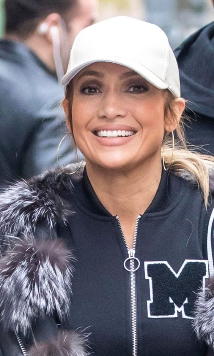 Jennifer Lopez walking in the street wearing a white baseball hat and a hoodie.