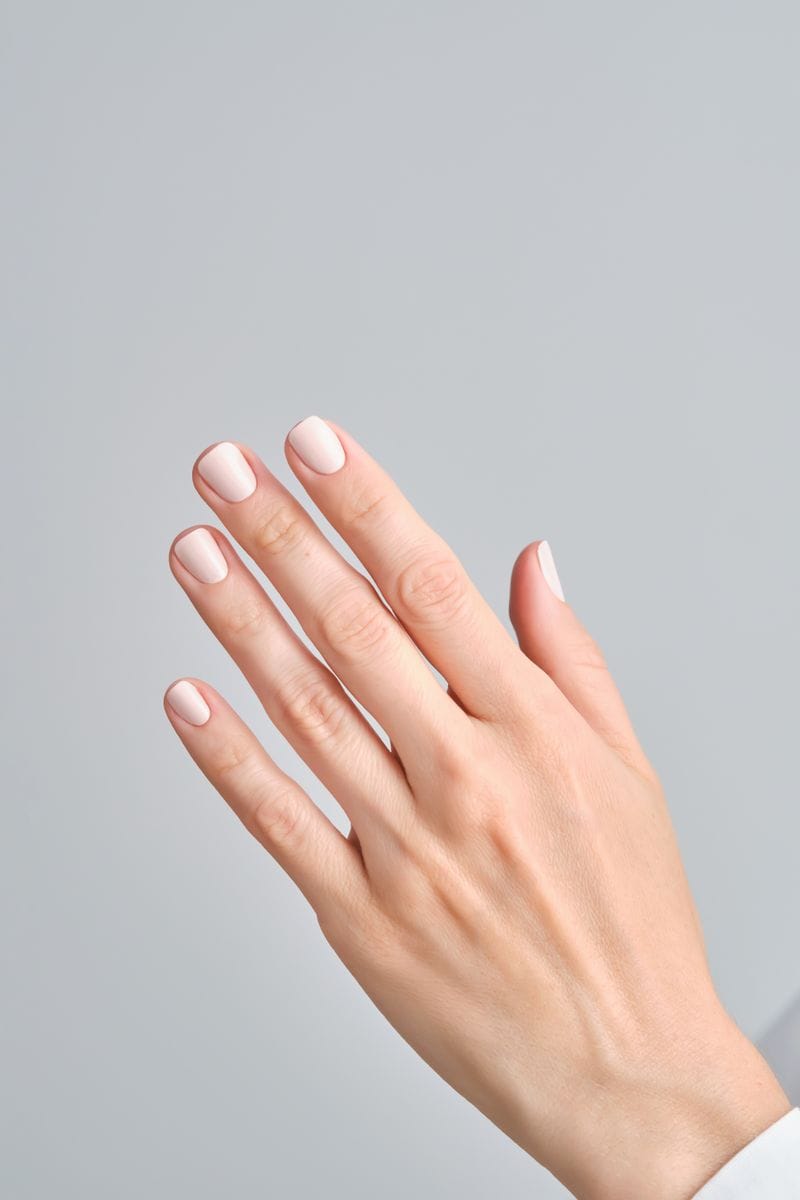 Close-up of a hand with short, neatly manicured nails painted in a soft nude shade against a neutral background.