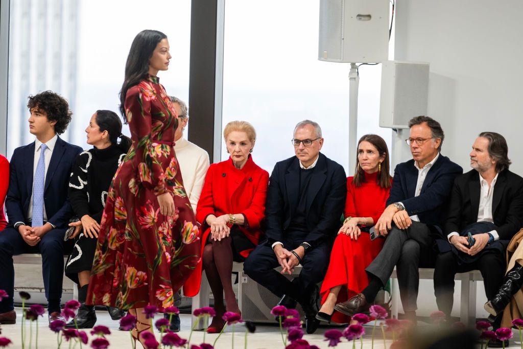 Carolina Herrera and other front-row guests observe a model in a red floral dress walking the runway at the Carolina Herrera Fall/Winter 2025 fashion show.