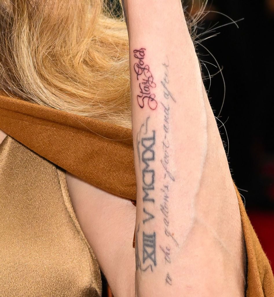 Close-up of Angelina Jolie's arm with multiple tattoos, including "Stay Gold" in red cursive and Roman numerals with script writing.