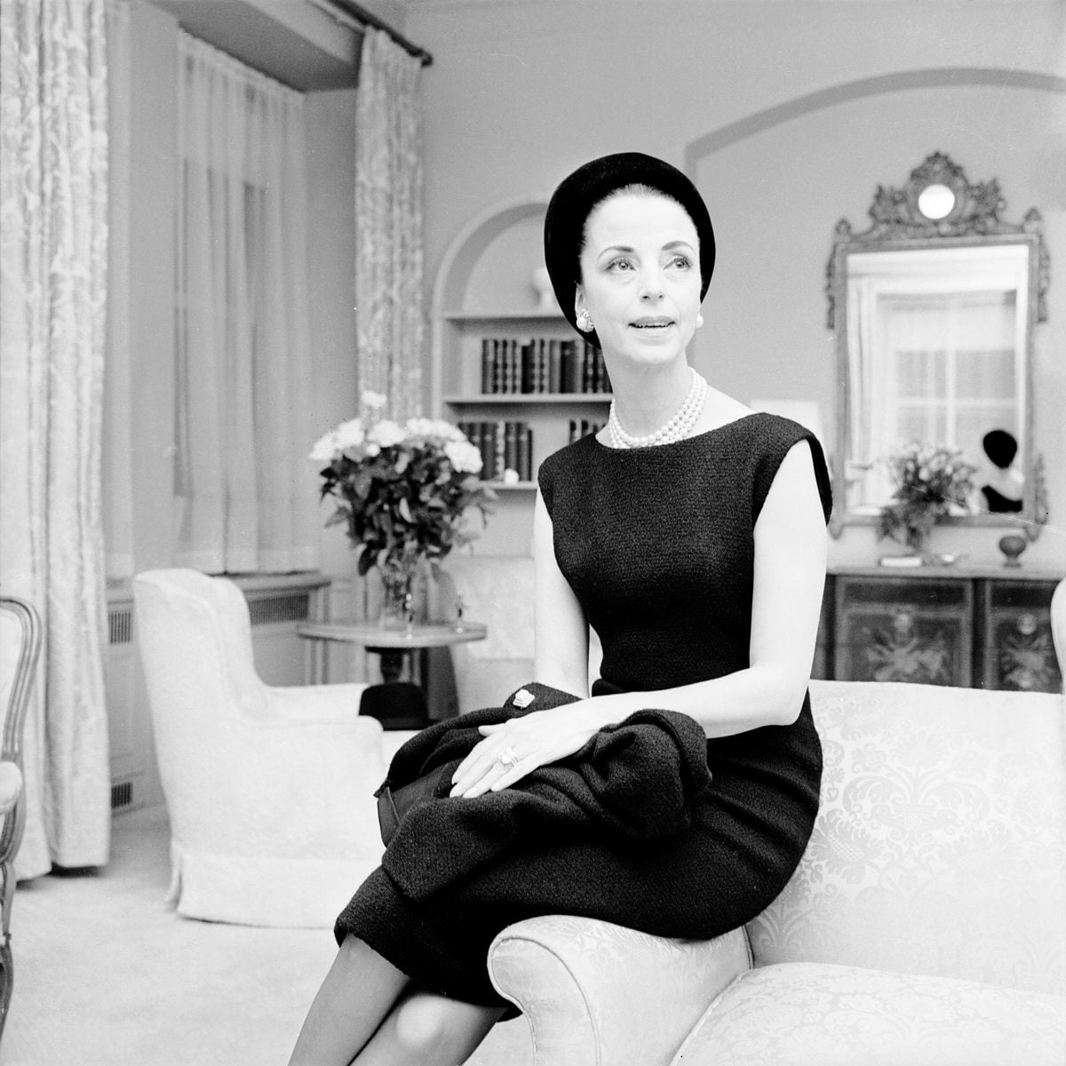 Gloria Guinness mengenakan jaket bulu hitam Givechy dengan pinggiran faille hitam di atas gaun tanpa lengan dengan topi Balenciaga hitam.