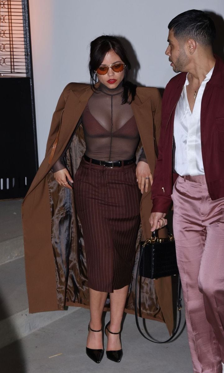 Jenna Ortega walking into a pre-Oscar event wearing a sheer brown turtleneck over a structured bralette, paired with a pinstriped midi skirt and a long brown overcoat. She completes the look with black pointed-toe heels, tinted sunglasses, and bold red lipstick, exuding a mix of vintage sophistication and modern edge.