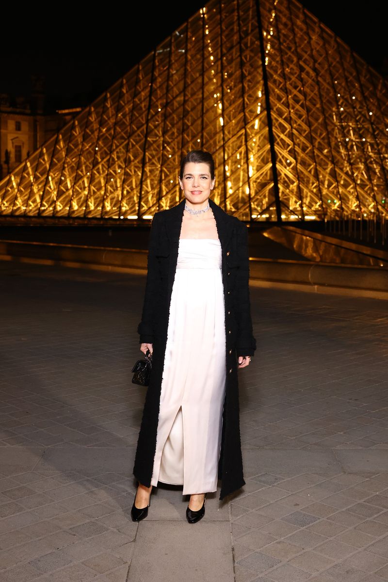 Charlotte Casiraghi poses in front of the illuminated Louvre Pyramid at night, wearing a strapless white gown, a long black coat, black heels, and carrying a small black handbag.