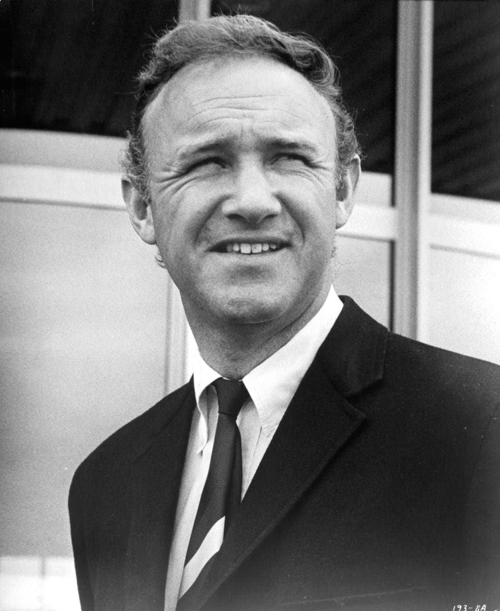 Black-and-white portrait of Gene Hackman in a suit and tie, smiling slightly while looking off to the side.