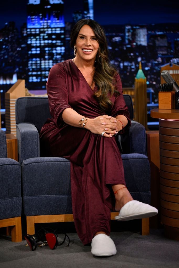 Karla Sofía Gascón smiling while seated on a late-night talk show set, wearing a burgundy satin dress and white fluffy slippers, with high heels on the floor beside her.