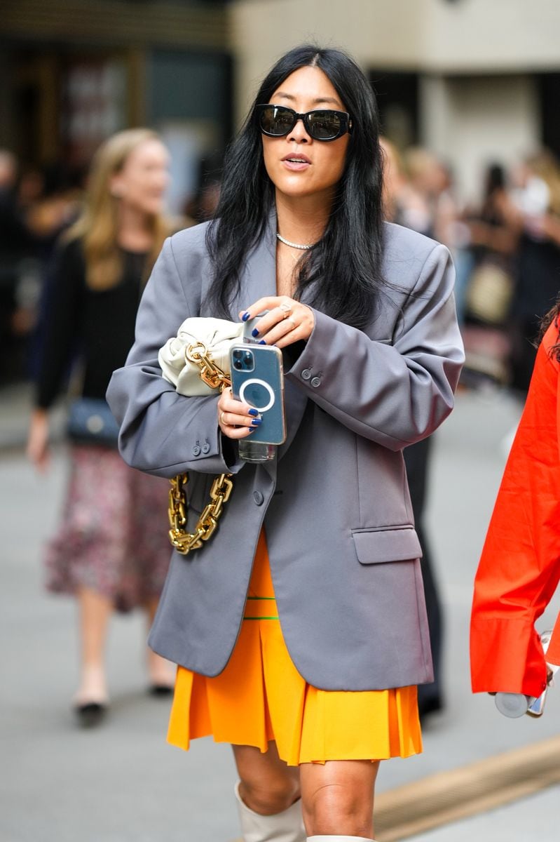 NEW YORK, NEW YORK - SEPTEMBER 13: A guest wears black sunglasses, a rhinestones necklace, a gray oversized blazer jacket, a white latte matte leather puffy handbag from Bottega Veneta, an orange pleated / accordion short skirt, white leather heels knees boots , outside Brandon Maxwell, during New York Fashion Week, on September 13, 2022 in New York City. (Photo by Edward Berthelot/Getty Images)