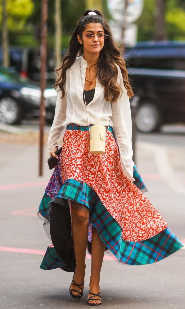 Noa Souffir wears sunglasses, a white shirt, a cylinder-shaped bag, a skirt with red floral print and green checked pattern