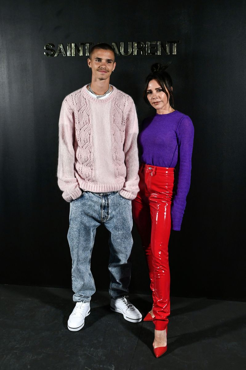 Romeo Beckham and Victoria Beckham posing together at a Saint Laurent event, standing against a black backdrop with the brand's logo. Romeo wears a pastel pink cable-knit sweater, acid-wash jeans, and white sneakers, while Victoria stuns in a purple fitted sweater and glossy red vinyl pants with matching heels.