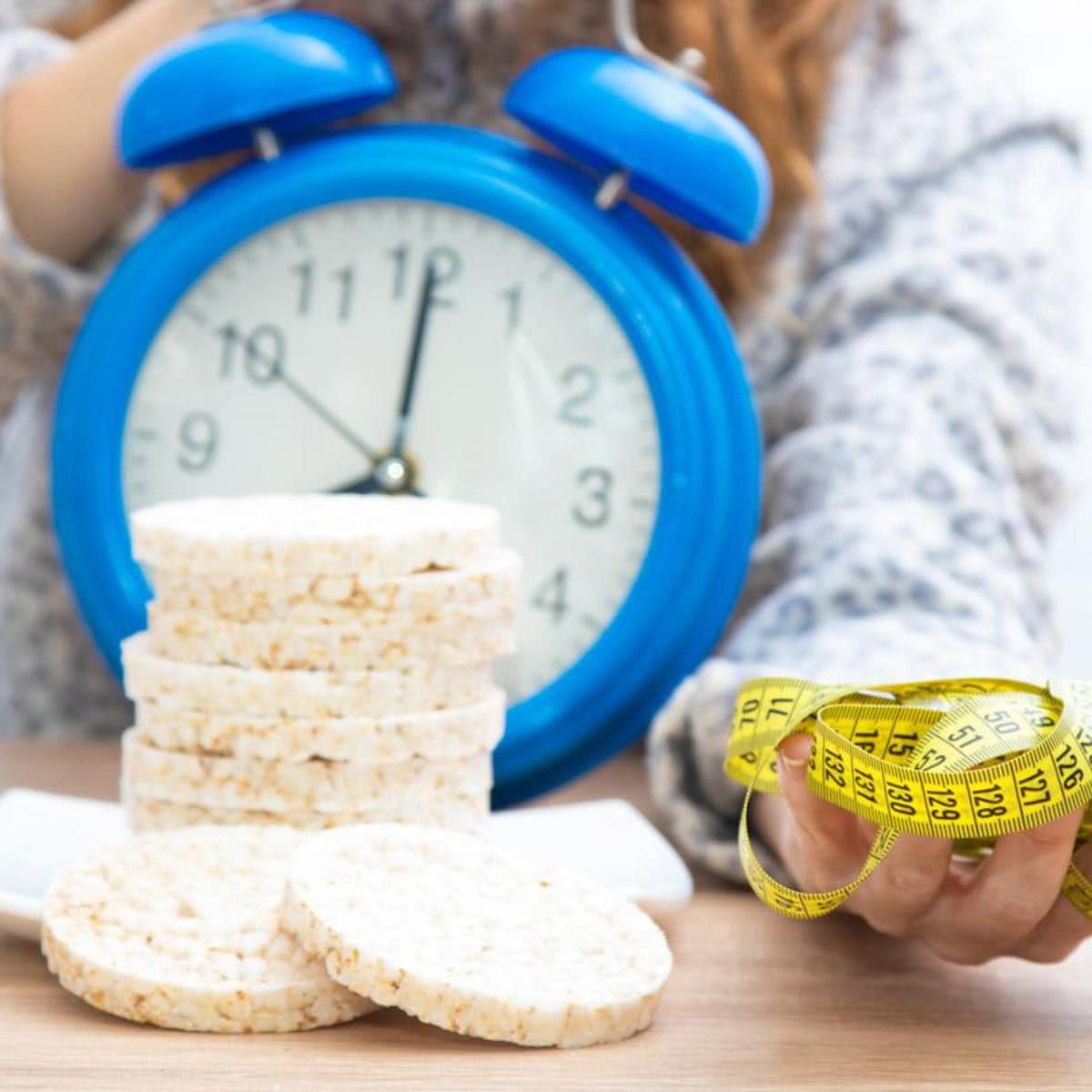 Mujer sostiene un reloj, junto a una cinta metrica y al lado galletas dieteticas