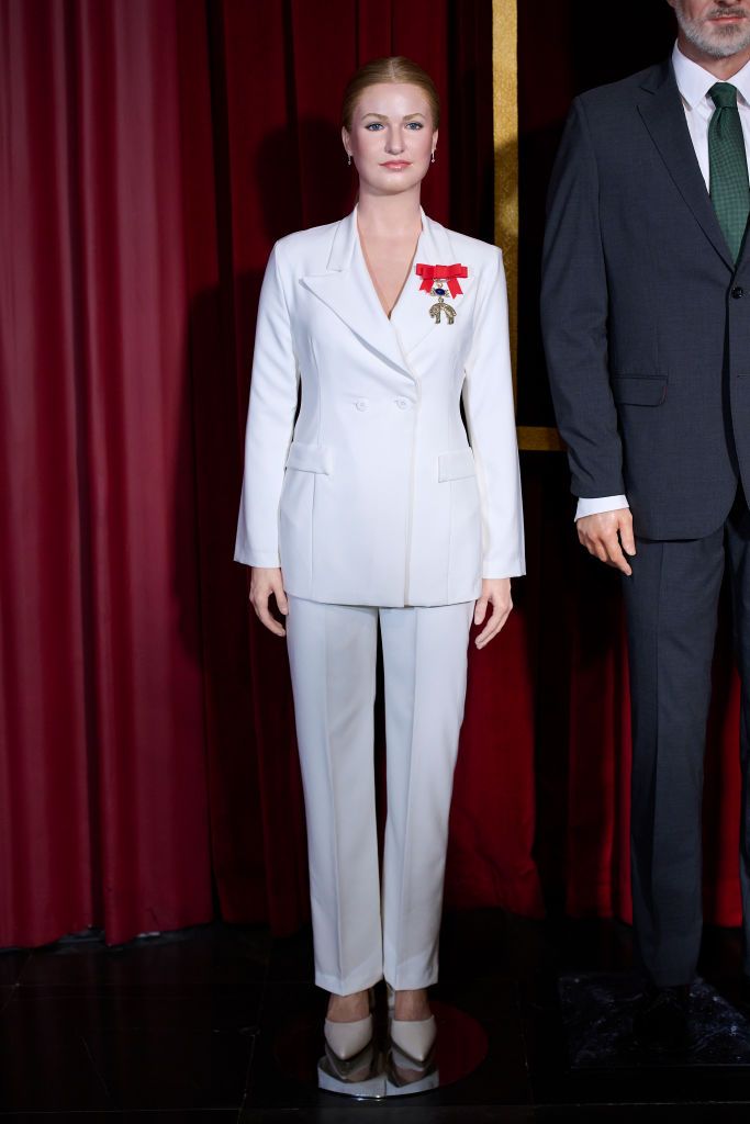 A full-body wax figure of Princess Leonor dressed in a white tailored suit with a red and gold medal, standing against a red curtain backdrop at the Madrid Wax Museum.