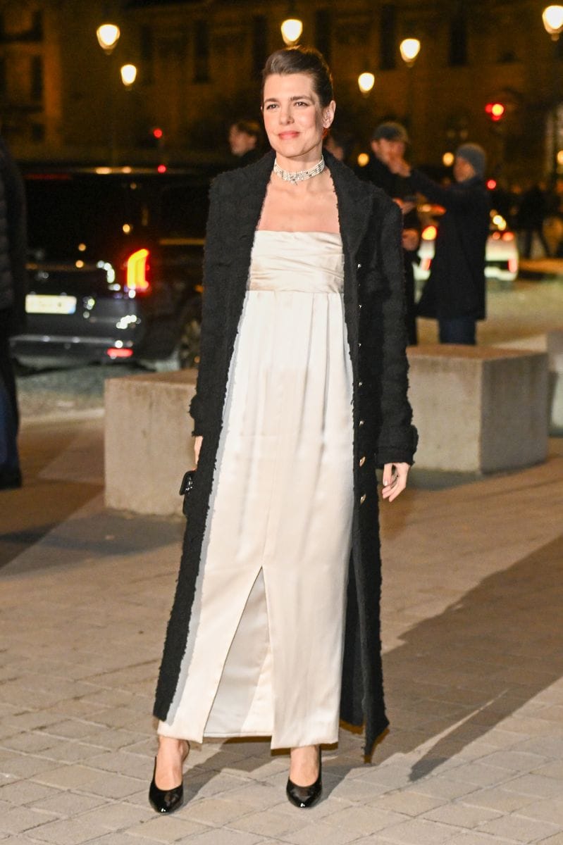 Charlotte Casiraghi poses in front of the illuminated Louvre Pyramid at night, wearing a strapless white gown, a long black coat, black heels, and carrying a small black handbag.