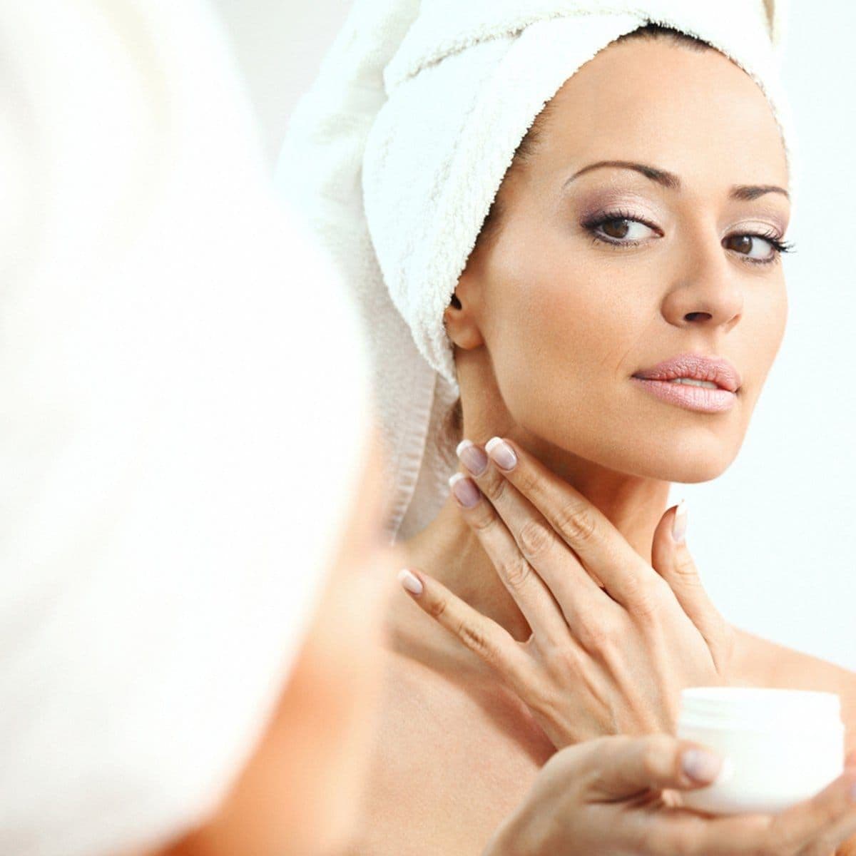 Mujer morena, con toalla blanca sobre su cabeza, se mira en un espejo y se aplica crema en el cuello