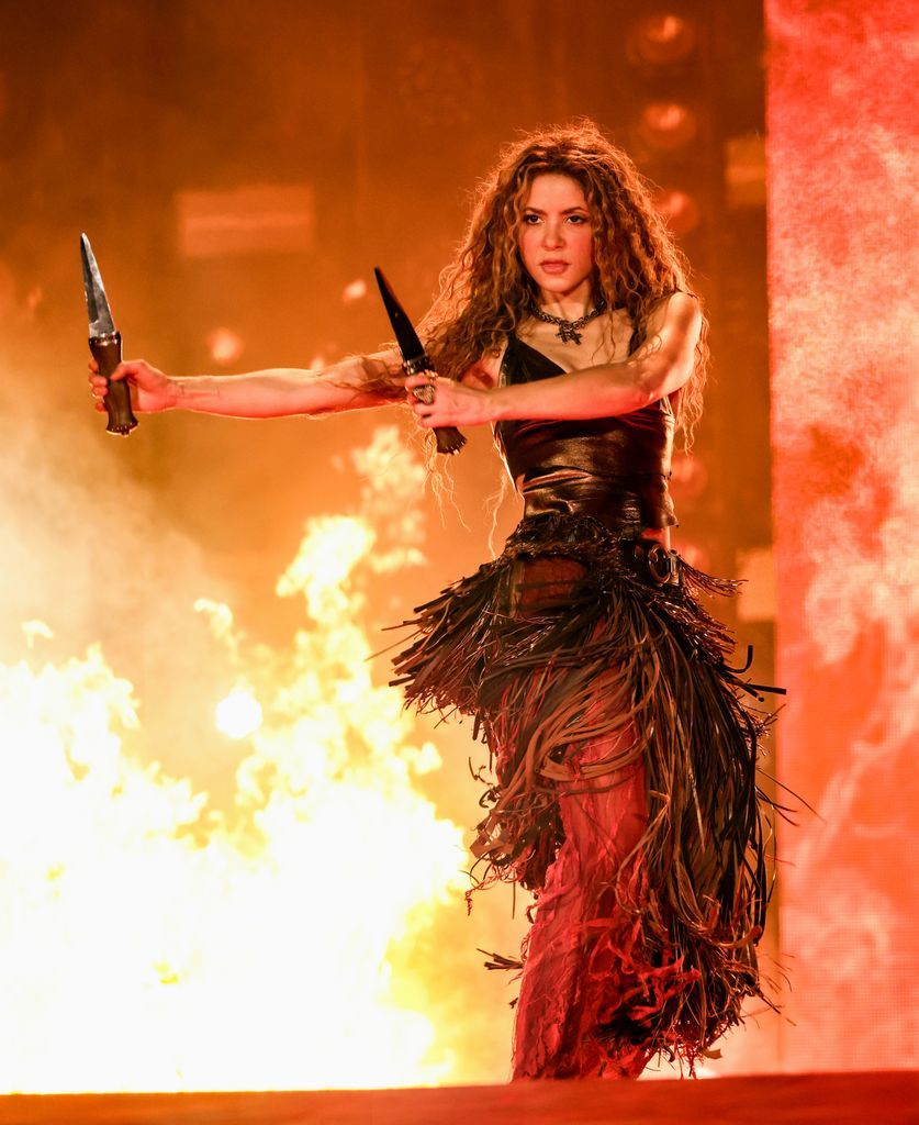 Shakira performs on stage in a warrior-inspired outfit, holding knives in both hands, with flames and fire effects in the background.