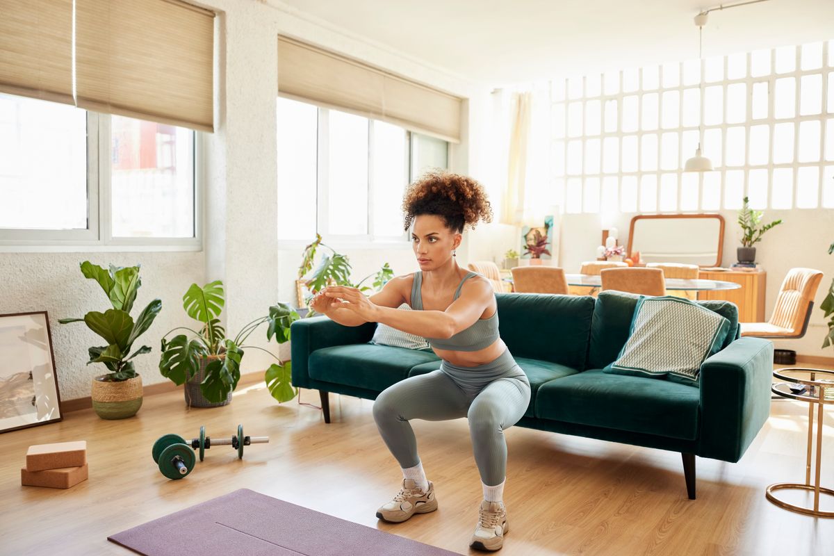 Full length of fit female doing squats in front of sofa. Young woman in sportswear is exercising at home. She is in living room. 