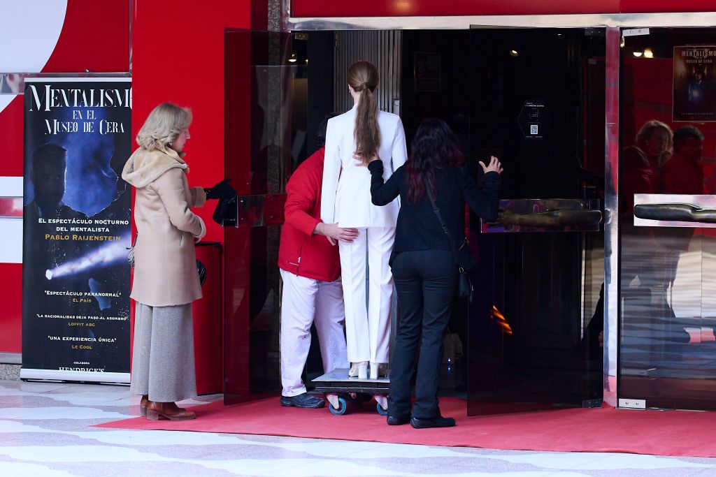 The wax figure of Princess Leonor, dressed in a white suit, being transported into the Madrid Wax Museum by staff members, with a red carpet and museum entrance visible.