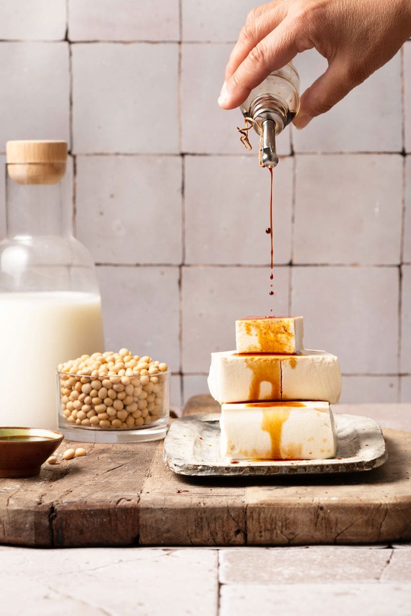 large group of plant based products on wooden board with copy space, soy milk in glass bottle, soy beans, tofu cubes, soy sauce and miso sauce, human hand pouring soy sauce on tofu cubes, copy space. 