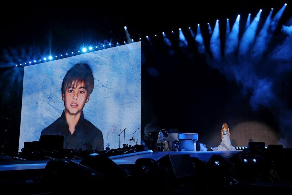 Shakira stands on stage near a piano, performing in front of a giant screen displaying a photo of her son, surrounded by blue lighting.