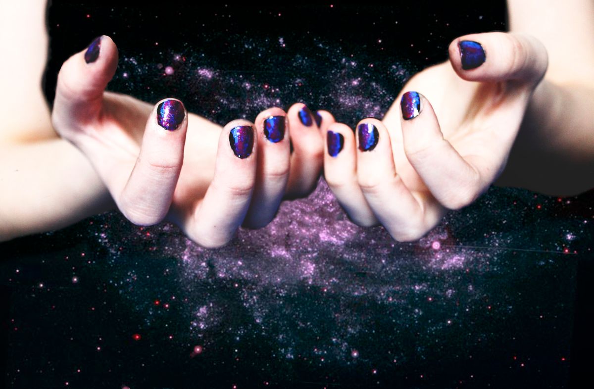 Hands with galaxy-themed nail art featuring shimmering purple, blue, and black tones, set against a cosmic background.