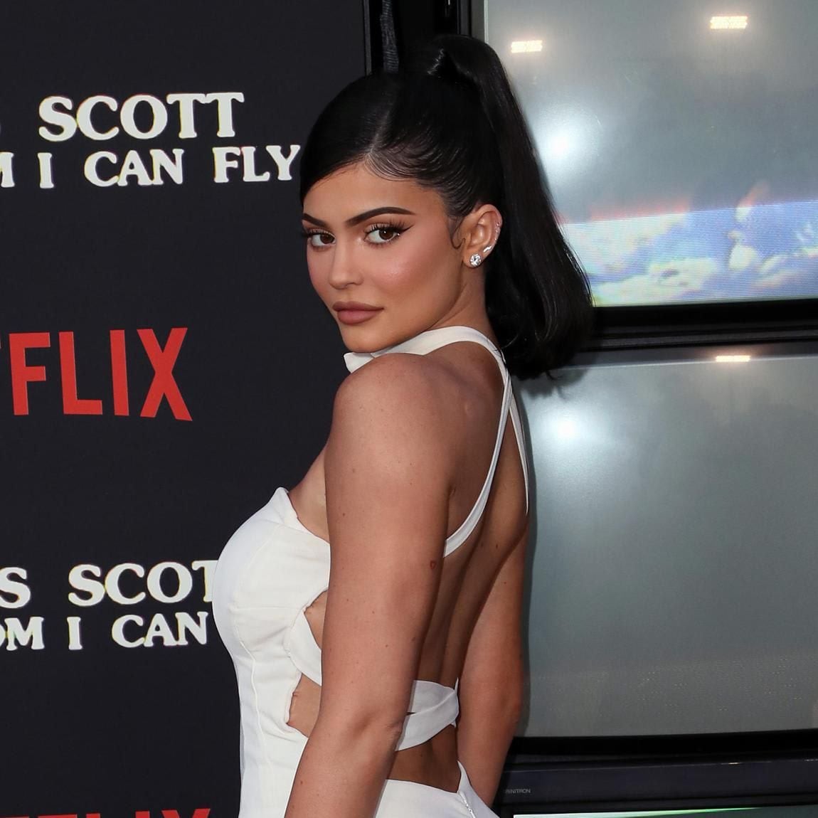 Kylie Jenner posing in profile showing off her dark ponytail wearing a white, open-back jumpsuit