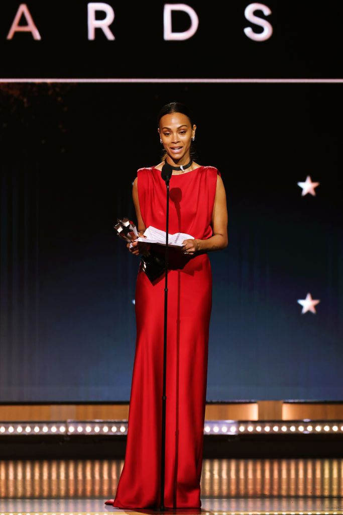 Zoe Saldaña in a red gown stands on stage delivering a speech while holding an award at the 30th Annual Critics Choice Awards.