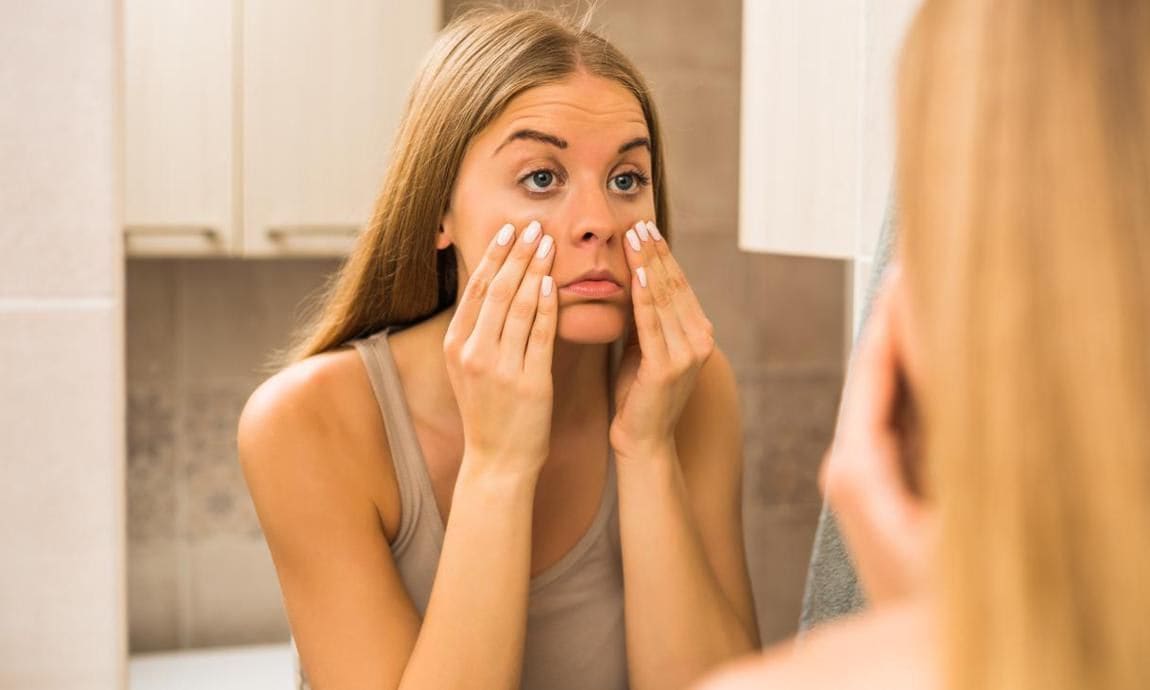 A woman looks at the bags under her eyes in a mirror