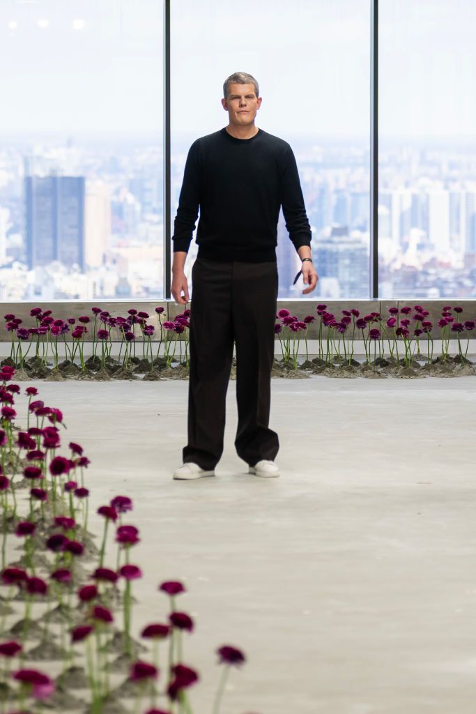 Wes Gordon standing on the Carolina Herrera Fall/Winter 2025 runway, with a cityscape view behind him and purple flowers lining the floor.