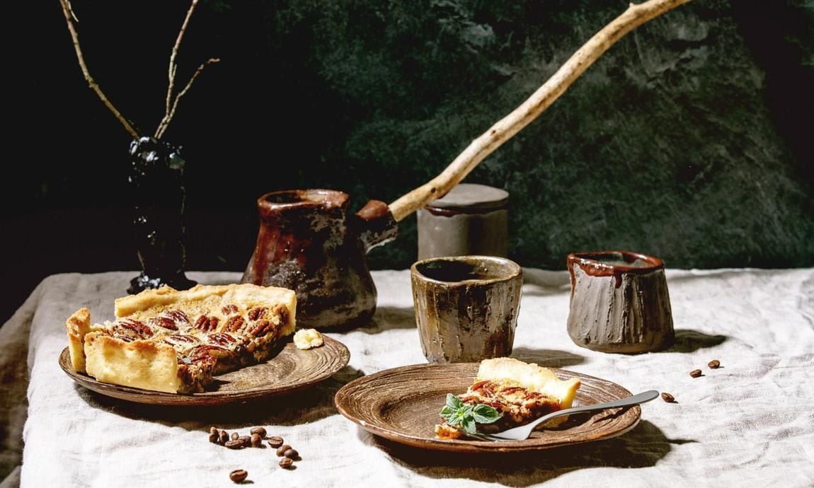 Piece of home baking classic american pecan caramel pie on ceramic plate, coffee cup, cezve, jug of cream on gray linen table cloth..