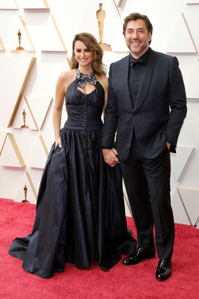 Penélope Cruz and Javier Bardem pose on the Oscars 2024 red carpet, both dressed elegantly in black, holding hands and smiling for the cameras