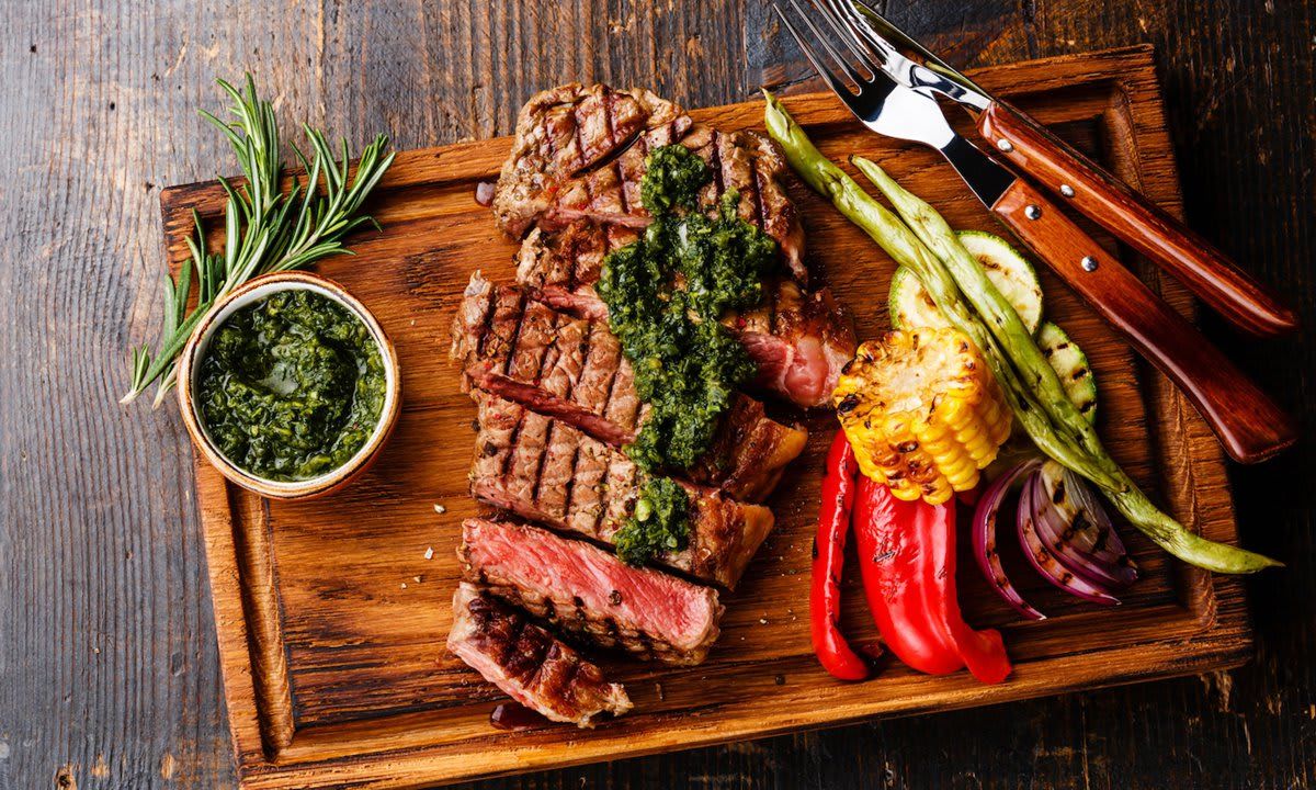 Sliced Striploin steak with chimichurri sauce and Grilled vegetables on cutting board on dark background