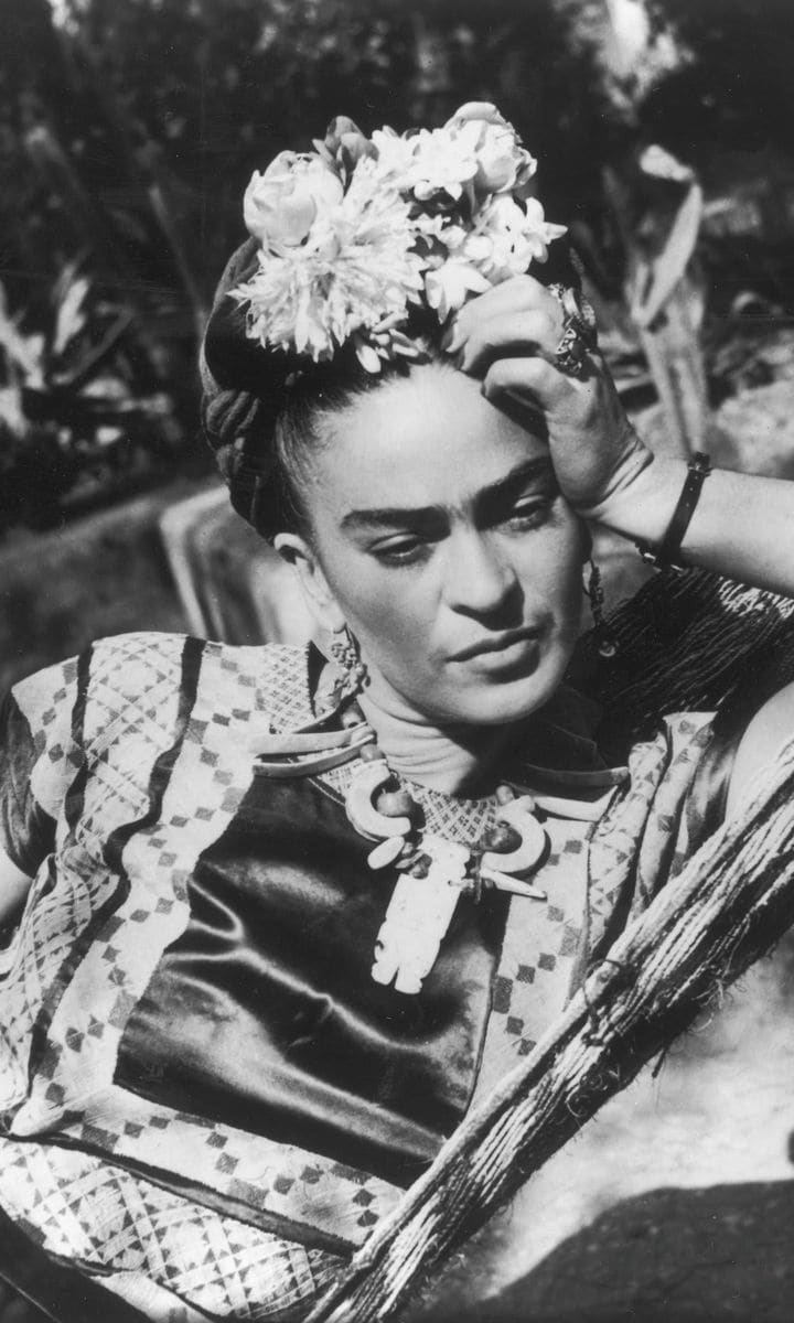 This is Kahlo wearing a folk costume and flowers in her hair, leaning her head on her hand while lying in a hammock.