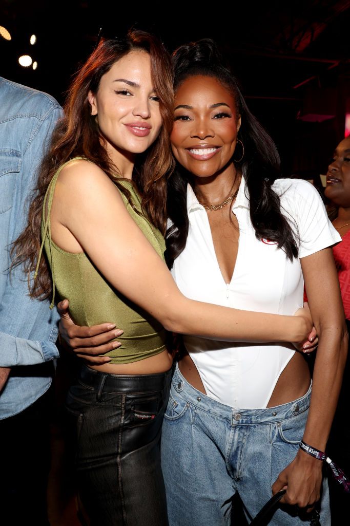 Eiza González in a green crop top hugging Gabrielle Union, who wears a white top and denim jeans at a Super Bowl event.