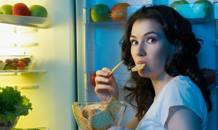 Woman eating crackers with the refrigerator door open and healthy food in the background