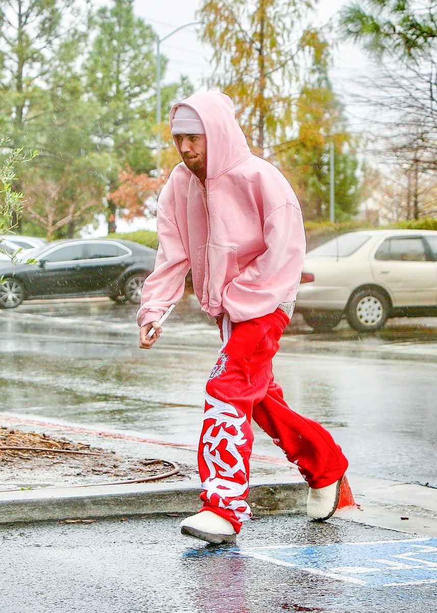 
Justin Bieber and a friend arrive at a go-kart track in Santa Clarita despite the rainy weather. The singer stands out in a light pink zip-up hoodie, a matching beanie, and bold red sweatpants with graffiti-style lettering. He completes the cozy streetwear look with fluffy white slip-on shoes. 