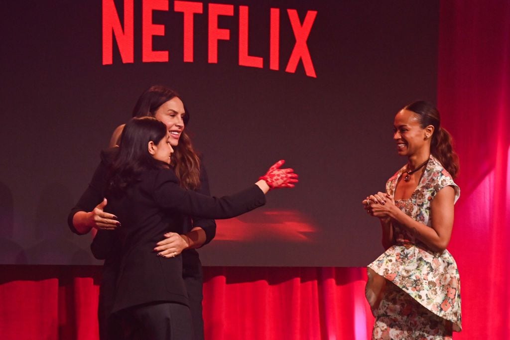 Karla Sofía Gascón and Zoe Saldaña on stage at a Netflix event, with Gascón embracing an attendee and Saldaña clapping, as the Netflix logo appears in the background.