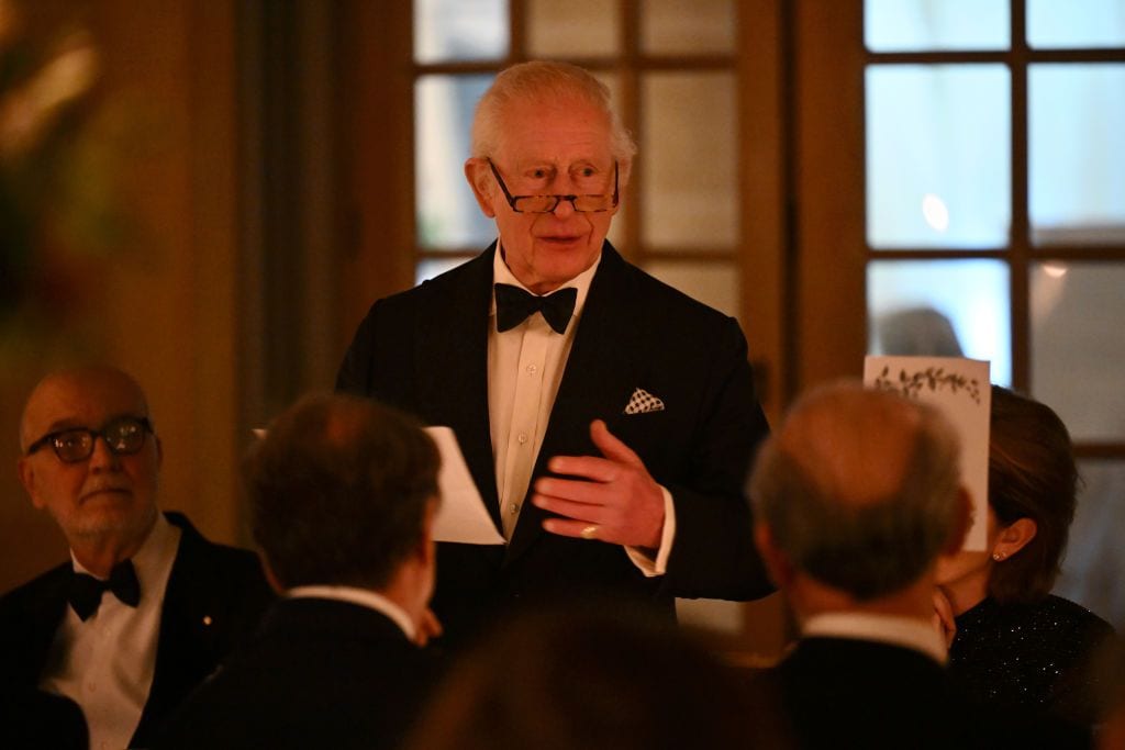 King Charles III delivers a speech at a formal gala dinner at Highgrove House, addressing distinguished guests in an elegant candlelit setting.