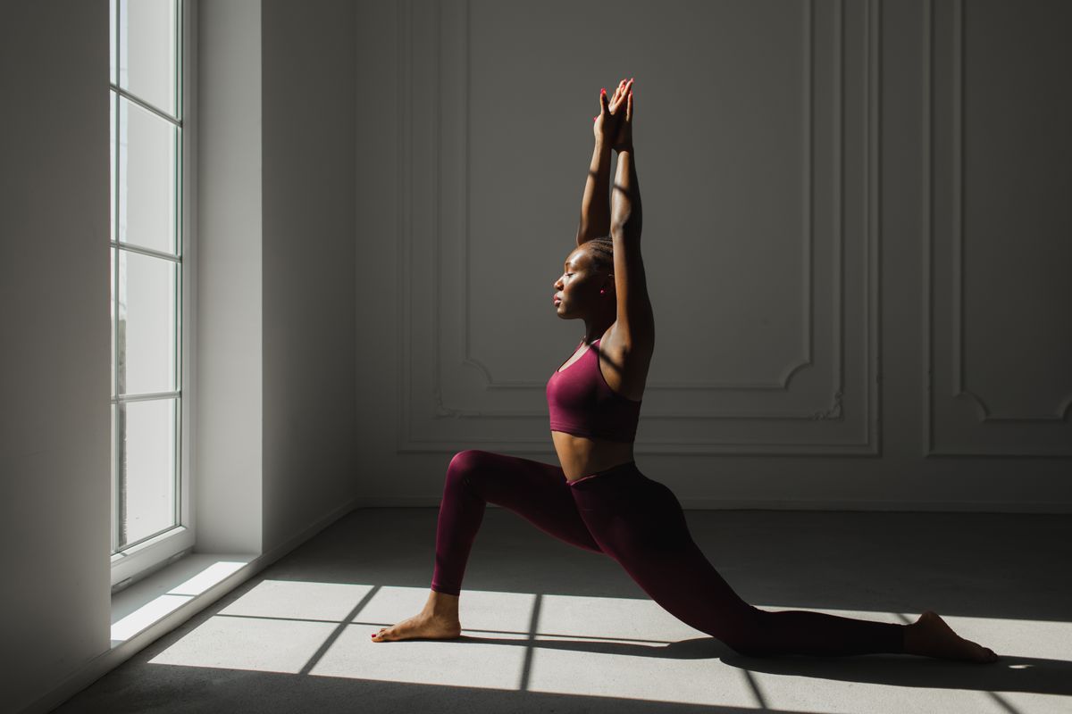 Fitness beautiful black woman with braided hair practicing yoga and doing variation of High Lunge, version of Virabhadrasana I (Warrior I Pose). Fit millennial African American female being in great shape, leading active lifestyle. Contrast light.