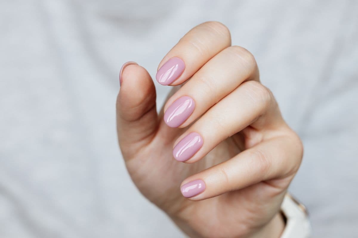 Close-up of a hand with short, glossy pastel pink nails against a neutral background.