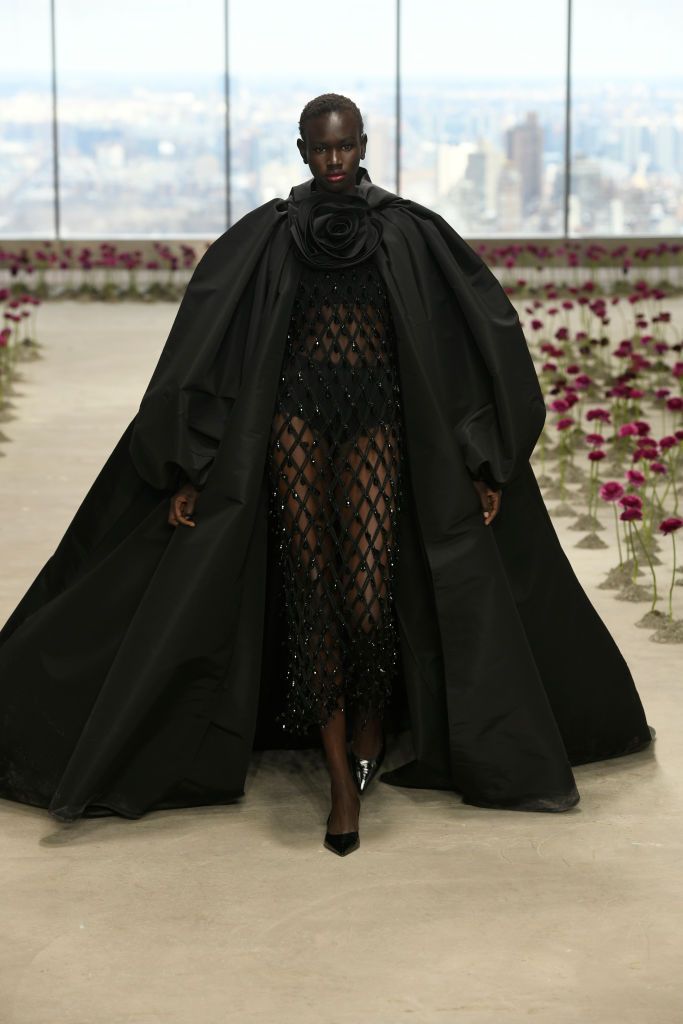 A model wearing a black oversized cape with a floral neckline and a sheer, embellished dress walks the Carolina Herrera Fall/Winter 2025 runway, with a cityscape in the background.