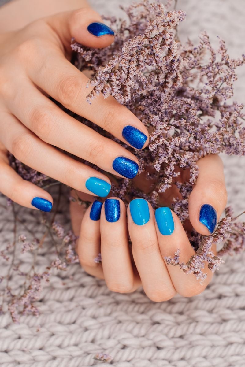 Close-up of hands with a blue manicure, combining glossy and glitter nail polish, holding dried lavender flowers on a knitted fabric background.