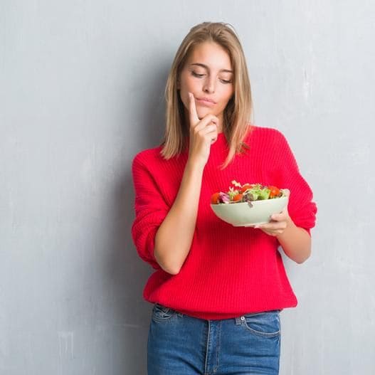 Woman in a red sweater, holding a bowl of food, looking doubtful about eating it