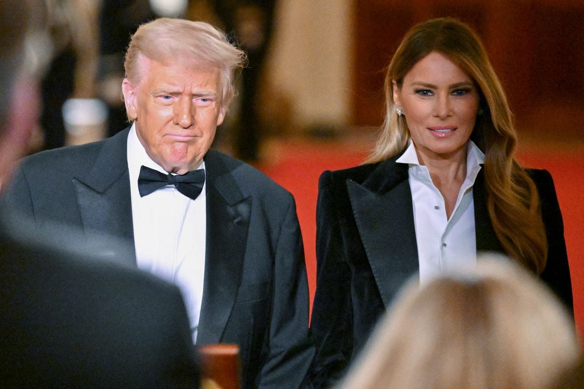 President Donald Trump and First Lady Melania Trump walk hand in hand on the red carpet at the White House, dressed in classic tuxedos, surrounded by uniformed personnel.